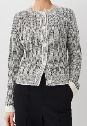 Kobieta ubrana w czarno-biały, teksturowany kardigan z dzianiny z białymi guzikami, na czarnych spodniach, ręce w kieszeniach, na jednolitym tle.