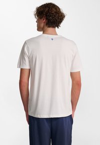 Vit bomull t-shirt med korta ärmar. Har en rund halsringning och en liten blå logotyp på baksidan nära halsringningen. Slät textur.