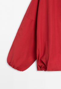 Red jacket sleeve with elastic cuff and visible side pocket seam on a plain light background.