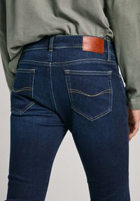 Temno modri jeans iz denima z rumenim šivom, z rjavim usnjenim oznako na hrbtu pasu in dvema zadnjima žepoma s ukrivljenimi šivi.