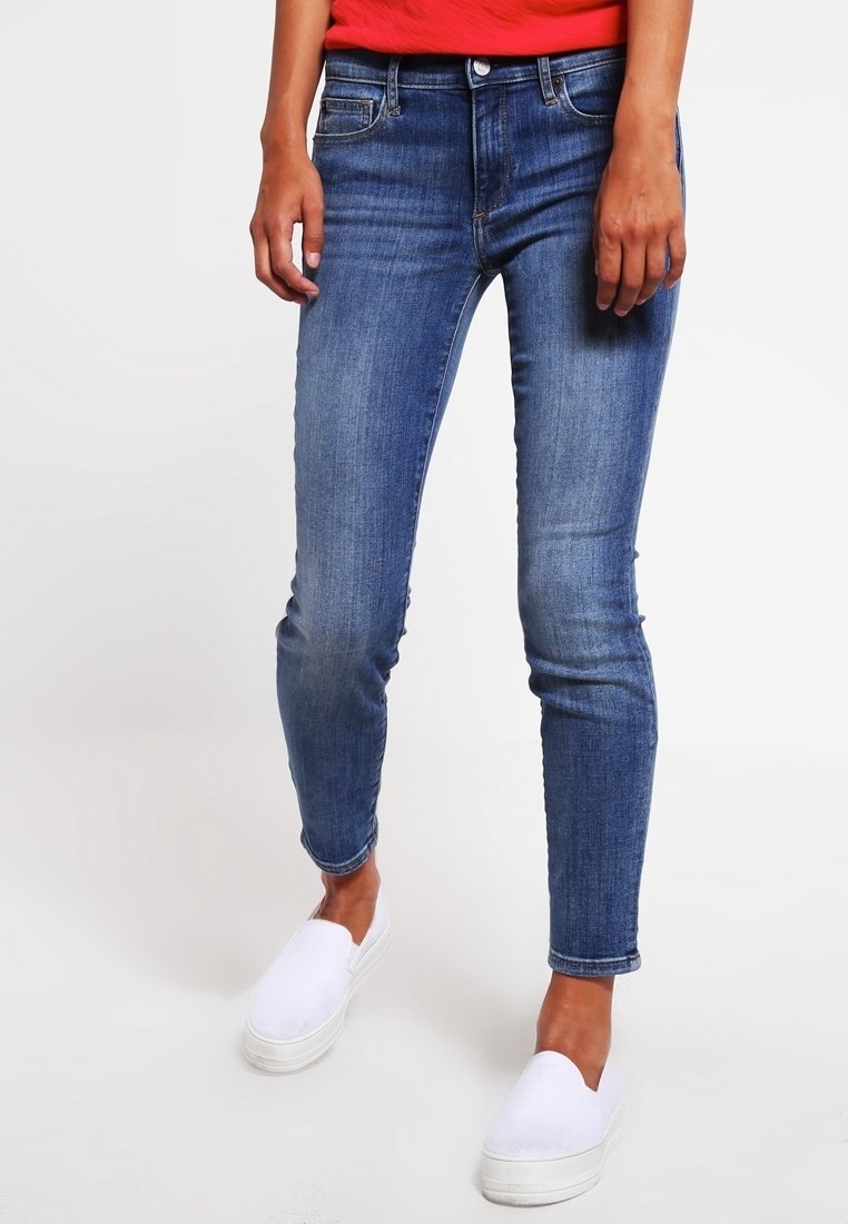 Skinny blue denim jeans with a faded finish, featuring five-pocket styling and a zip fly. Paired with white slip-on shoes.