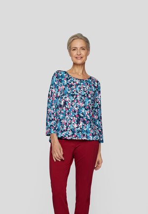 Bluse mit blauen Blumenmuster und 3/4-Ärmeln, verziert mit pinken und weißen Blumen, kombiniert mit roten Hosen. Glatte Textur, lässige Passform.