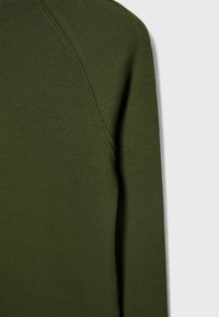 Gros plan sur un vêtement à manches longues côtelé de couleur vert olive, avec des coutures visibles le long de l’épaule et de la manche.