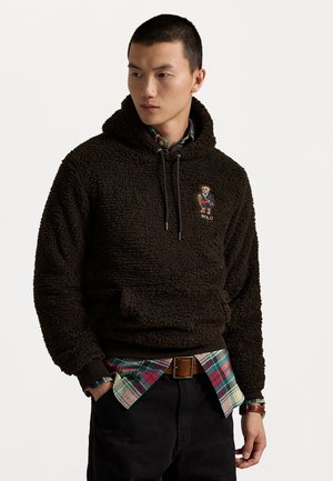 Felpa con cappuccio marrone in pile con finitura testurizzata, caratterizzata da un logo di orso ricamato. Indossata sopra una camicia a quadri, con pantaloni scuri e una cintura.