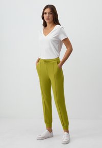 Weißes V-Ausschnitt-T-Shirt kombiniert mit leuchtend grünen Jogginghosen mit elastischen Bündchen und Seitentaschen, ergänzt durch weiße Sneakers.