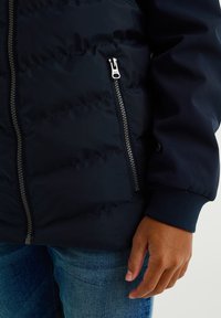 Navy puffer jacket featuring a zippered pocket, ribbed cuffs, and a quilted texture. Smooth fabric with durable hardware details.