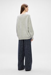 Suéter cinza de malha com um corte descontraído e punhos canelados, emparelhado com calças largas de tecido texturizado em azul-marinho. Detalhe notável: pequeno botão nas costas.