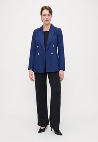 Sini kaksirivinen bleiseri kultanappien kanssa, yhdistetty mustiin teksturoituihin leveälahkeisiin housuihin ja teräväkärkisiin mustiin korkokenkiin.