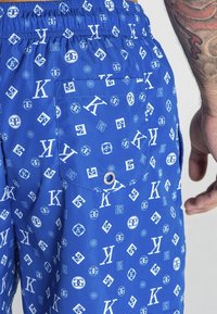 Blå badshorts i ett lätt material har en flerfärgad logotryck i vitt. Inkluderar en bakficka med en ögla.