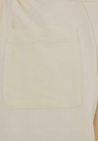 Tissu beige avec une poche avant rectangulaire cousue sur un vêtement, montrant un gros plan de la poche et du matériau environnant.