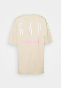 Camiseta de algodón beige de manga corta, con gran texto blanco "GAP" y estampado rosa "Original" en la parte trasera. Diseño sencillo, ajuste relajado.