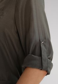 Chemise en tissu léger olive, avec des manches retroussées fixées par des boutons et un léger éclat ; présente une texture douce et une coupe ajustée.