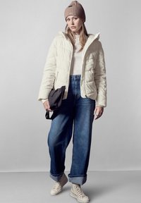 Giacca trapuntata in bianco sporco, con una consistenza spessa, abbinata a jeans blu a vita alta e sneaker in tela beige. Accentata da una borsa nera.