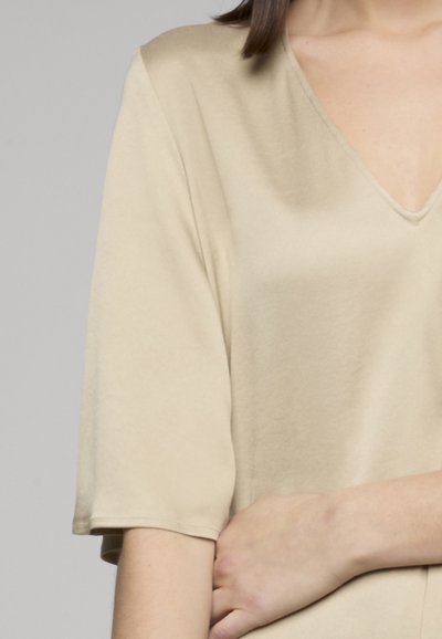 Personne portant un t-shirt beige à manches courtes en V, avec un bras croisé et la main posée sur le coude opposé.
