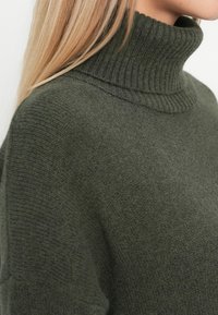 Temna zelena pulover s turtleneckom, ribanim ovratnikom in teksturiranim pletenim vzorcem, ki prikazuje mehak material in sproščen kroj.
