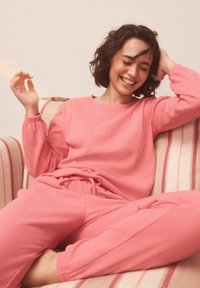 Ružová pletená sada na doma s dlhými rukávmi a elastickým pásom; nohavice vo voľnom strihu, textúrovaná látka, osoba sedí na pruhovanej pohovke.