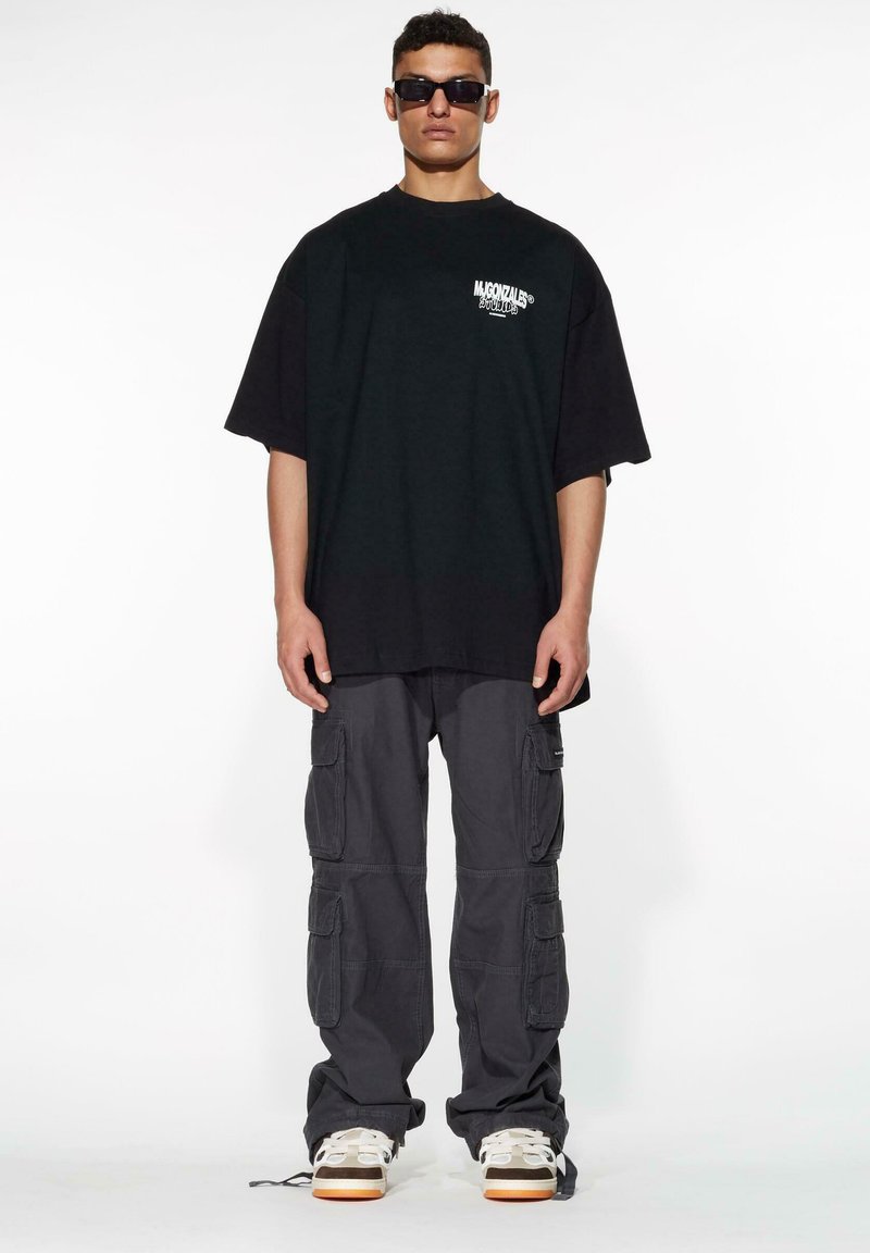 Svart oversized t-skjorte med hvit logo, kombinert med grå cargo-bukser med flere lommer og lyse sneakers med hvite detaljer.
