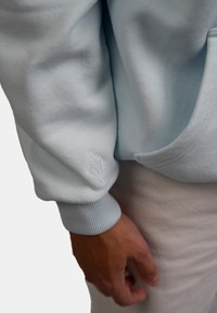 Felpa blu chiaro con polsini a costine e un dettaglio ricamato bianco a forma di diamante sulla manica. Tessuto morbido e textured visibile.