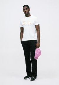 Vīrietis ģērbts baltā Versace Jeans Couture T-kreklā, melnās biksēs un melnos sporta apavos, labajā rokā tur rozā Versace Couture cepuri.