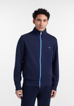 Ung mand iført en marineblå jakke med lynlås i lyseblå og en lille hundelogo på brystet, stående foran en enkel hvid baggrund.