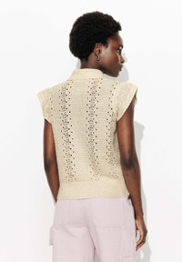 Femme aux cheveux courts et bouclés, portant un haut en tricot sans manches beige avec des trous décoratifs et un pantalon rayé rose clair, de profil.