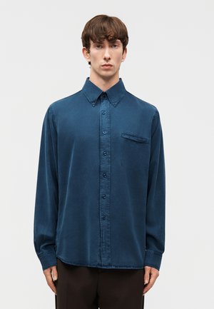 Tânăr purtând o cămașă închisă la culoare, cu nasturi și buzunar la piept, stând în fața unui fundal simplu, deschis la culoare.