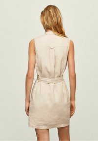 Donna con capelli biondi che indossa un vestito di lino beige senza maniche con cintura, in piedi di spalle contro uno sfondo chiaro e uniforme.