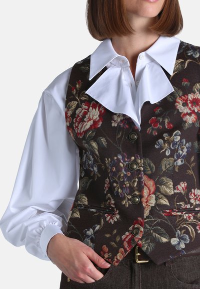 Femme portant un chemisier blanc à manches longues avec un large col et un gilet boutonné à motif floral foncé, main reposant dans la poche.