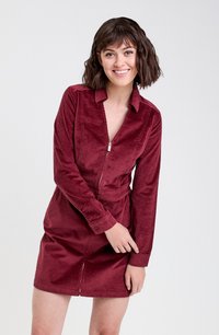 Robe en velours côtelé bordeaux avec manches longues, col et fermeture éclair à l'avant. Présente une texture à côtes verticales et une taille cintrée avec une jupe évasée.