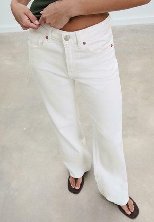 Osoba zakopčava gumb na bijelim trapericama visokog struka s širokim nogavicama, nosi tamnu majicu i crne japanke na betonskom podu.