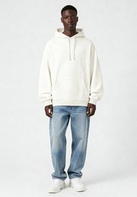 Felpa bianca in cotone con tasca frontale, abbinata a jeans azzurri e sneakers bianche. Vestibilità comoda, maniche lunghe e cappuccio regolabile con cordino.