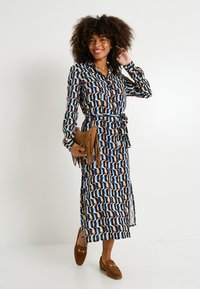 Vestido de manga larga, con cinturón, en un patrón geométrico multicolor que presenta tonos azules, marrones y crema. Combinado con un bolso de mano beige con flecos y zapatos marrones.