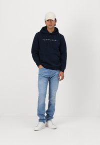 Marineblauwe hoodie met logo, lichtblauwe spijkerbroek en witte sneakers. Het model draagt een beige baseballcap. Algeheel casual outfit tegen een witte achtergrond.
