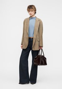 Νεαρό άτομο φοράει μπεζ σακάκι, ανοιχτό μπλε τοπ, φαρδιά σκούρα παντελόνια, κρατώντας σκούρα καφέ δερμάτινη τσάντα, στέκεται μπροστά σε απλό φόντο.