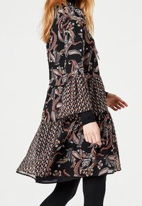 Černé šaty s paisley vzorem v oranžové, modré a bílé barvě. Mají vrstvený design s širokými rukávy a černými manžetovými detaily.