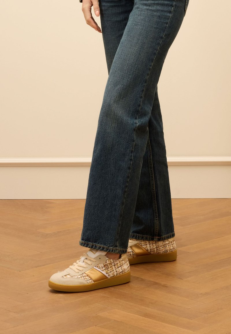 Sneaker mit beigen und goldenen Akzenten, strukturiertem Stoff und einem gemusterten Seitenbereich; kombiniert mit dunkelblauen Schlagjeans auf einem Holzboden.