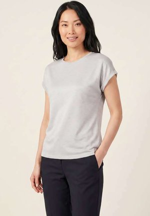 Mujer con cabello largo y negro que lleva una camiseta de manga corta gris claro y pantalones negros, sonriendo con una mano en el bolsillo.