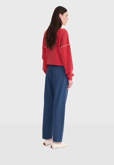 Femme aux longs cheveux noirs portant un pull rouge ample, un pantalon bleu taille haute et des chaussures blanches, debout de profil sur un fond uni.