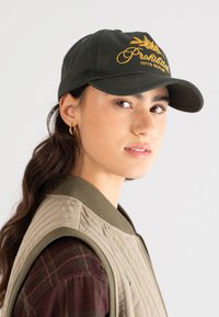 Young woman wearing olive green cap with yellow embroidery, gold hoop earrings, plaid shirt, and quilted vest, looking slightly sideways.