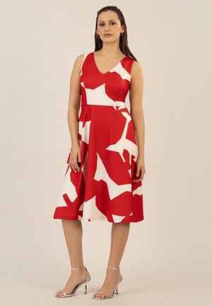 Rotes Kleid mit weißen abstrakten Mustern, V-Ausschnitt, ärmelloses Design, ausgestellt Rock, aus glattem Stoff. Das Model trägt silberne Riemchenschuhe.