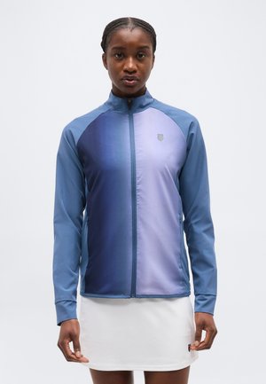 Nainen katsoo eteenpäin, yllään sinivioletti liukuvärinen vetoketjullinen takki ja valkoinen hame yksiväristä taustaa vasten.