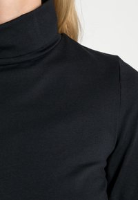 Top de cuello alto negro con un diseño ajustado, textura suave y tejido acanalado. El cuello es alto y estructurado, realzando la silueta.