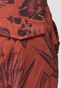 Tissu couleur rouille avec imprimés floraux et feuilles sombres, doté d'une poche à rabat boutonnée et d'une petite étiquette métallique de la marque.