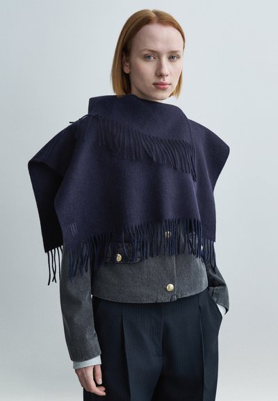 Νεαρή γυναίκα με κόκκινα μαλλιά που φοράει μπλε μάλλινο πόντσο με φουντίτσες πάνω από γκρι μπουφάν και σκούρο παντελόνι με ρίγες, σε απλό φόντο.