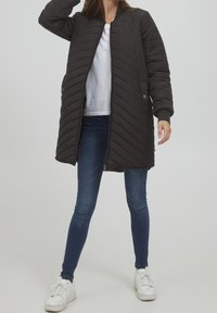 Svart quiltad lång jacka med blixtlås framtill, ribbade ärmslut och sidofickor, bärs över en vit skjorta och blå jeans, ihop med vita sneakers.