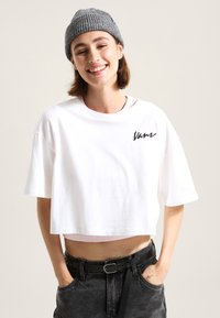 Camiseta corta blanca con mangas cortas y logo de "Vans" en negro. Combinada con jeans oscuros de cintura alta y un gorro de punto gris.