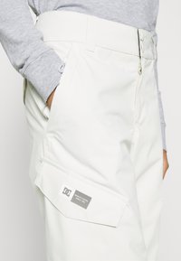 Witte cargobroek met een gladde textuur, voorzien van een zijzak en een zichtbaar logo label. Knop- en ritssluiting bij de taille.