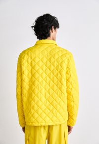 Casaco amarelo acolchoado com colarinho, apresentando um padrão de diamante e um corte solto. O modelo também usa calças amarelas combinando.