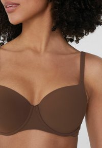 Bruine gepolsterde beha met gemoldeerde cups, verstelbare bandjes en een gladde textuur. Heeft gestikte details in het midden en een zachte stofafwerking.