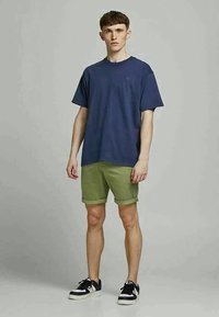 T-shirt in cotone blu navy abbinata a pantaloni corti verde chiaro. I pantaloni corti presentano orli arrotolati. Il modello indossa sneakers nere. Sfondo semplice.